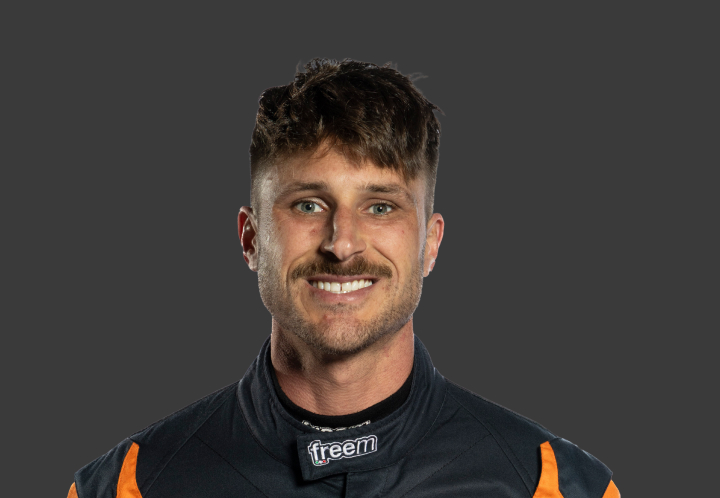 A headshot of professional T G R A driver Craig Thornton in a black and orange racing suit against a grey background.