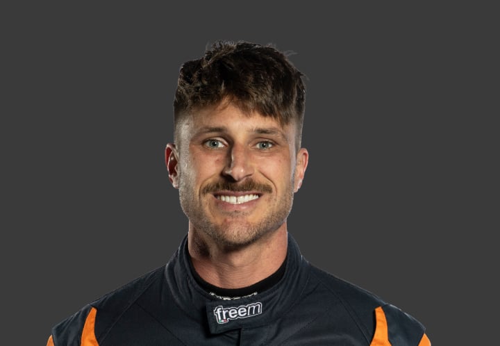 A headshot of professional T G R A driver Craig Thornton in a black and orange racing suit against a grey background.