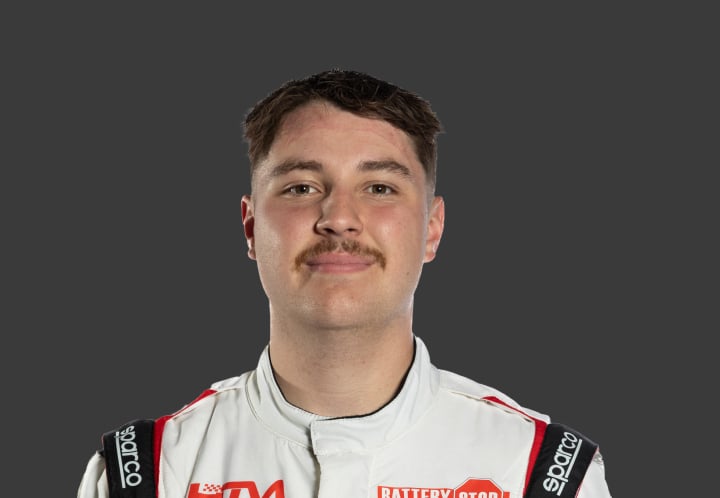A headshot of professional T G R A driver Harry Tomkins in a white, black and red racing suit against a grey background.