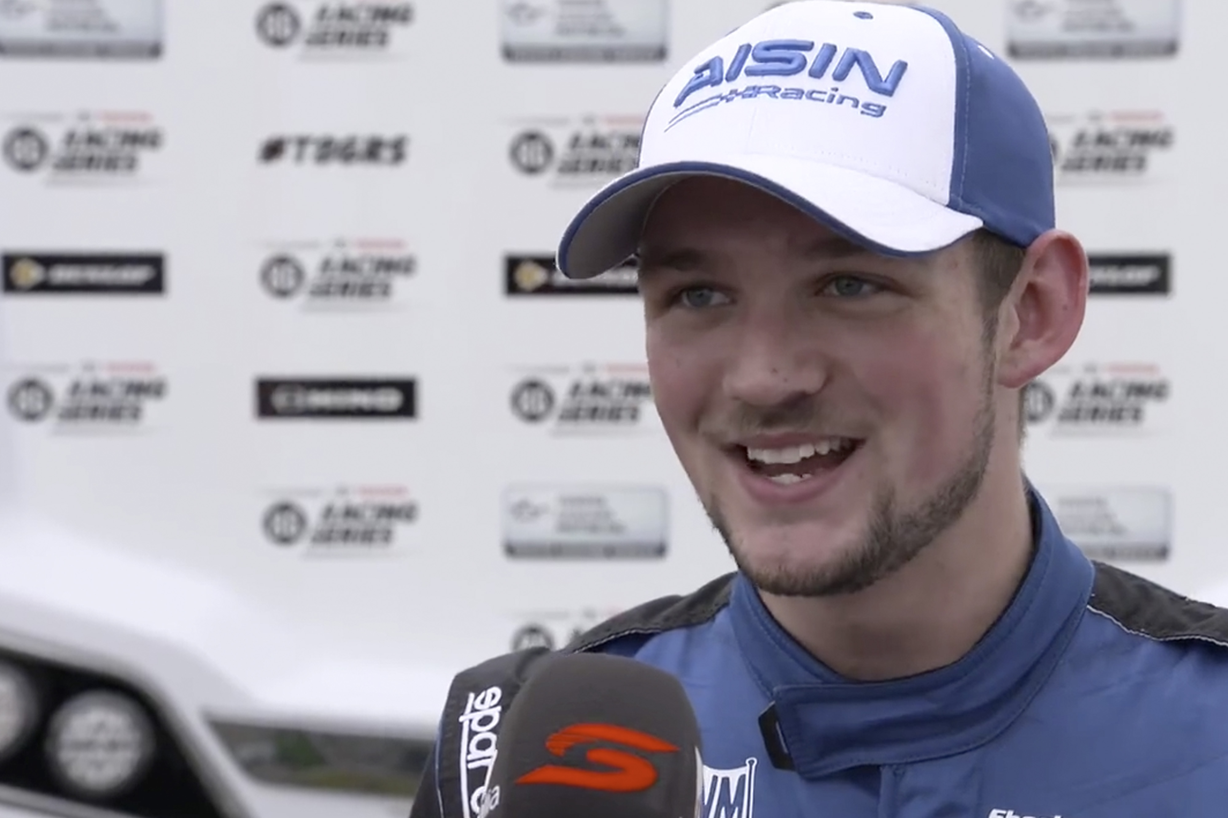 Professional racing driver Luke King smiles during an interview in front of a white media wall. He wears a blue and white cap and a matching racing suit.