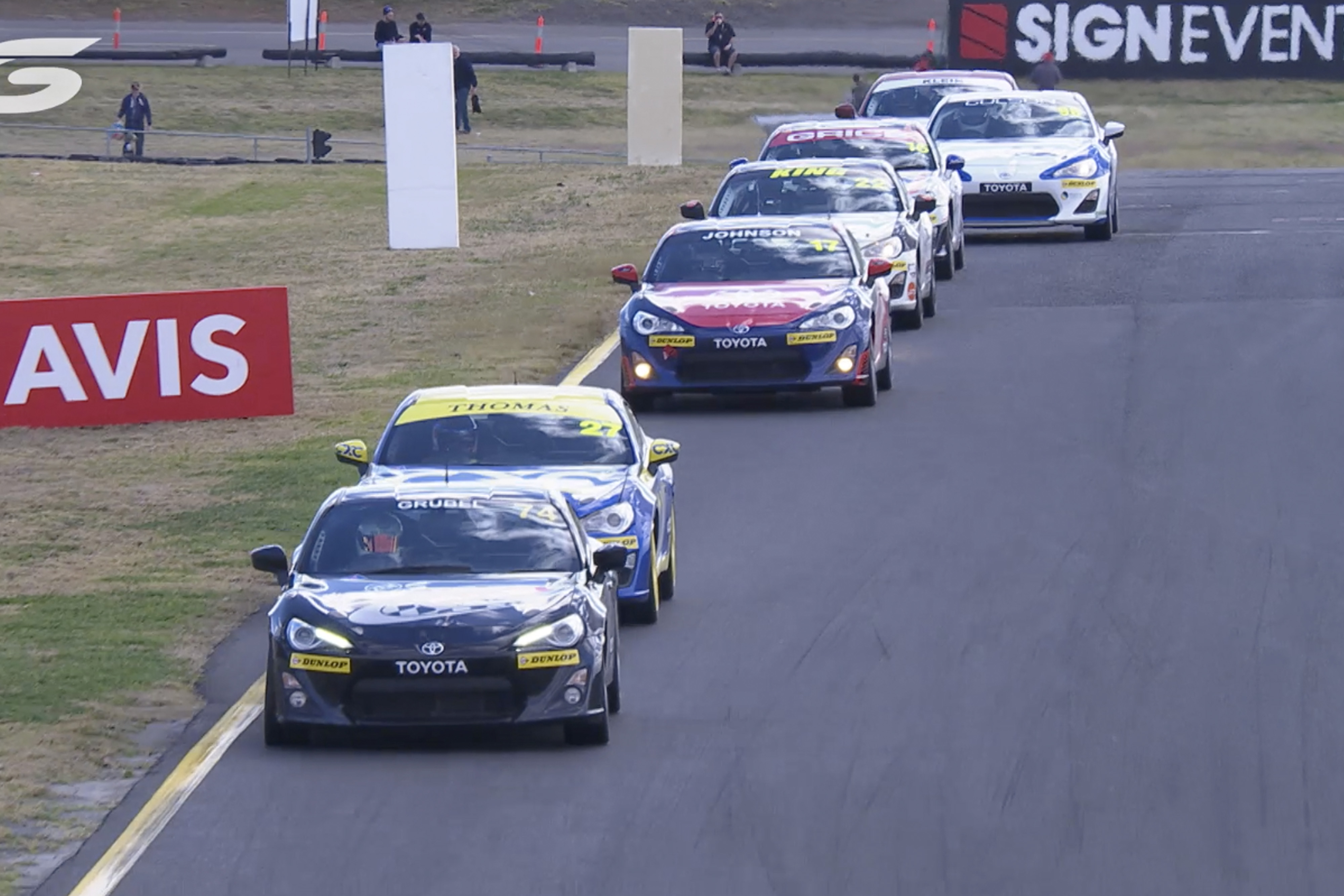 Toyota G R 86s in racing livery competing on a racetrack. They all race in a line towards the centremost side of the track.