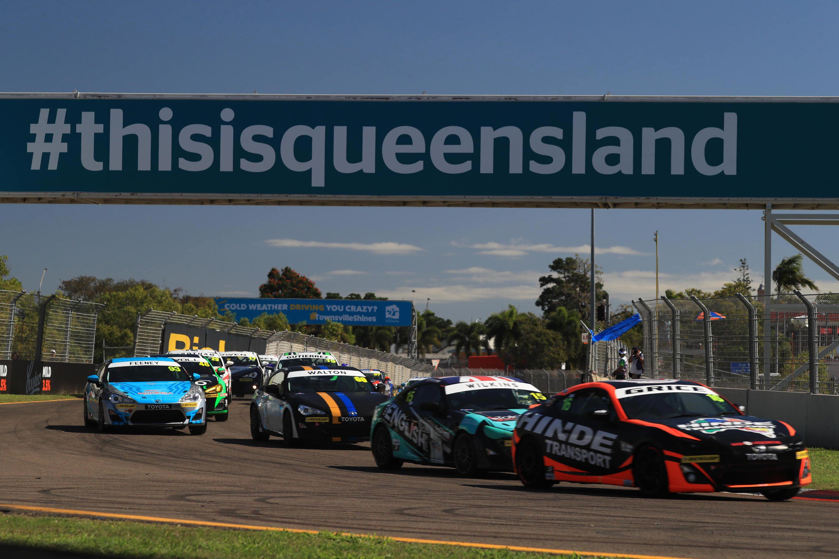 Toyota G R 86s in racing livery competing on a racetrack on a sunny day. A " hashtag this is queensland" banner is displayed over the track.