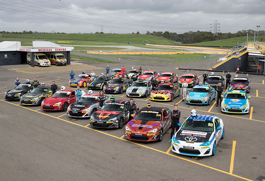 An aerial view of a parking lot full of Toyota G R 86s in racing livery, surrounded by green grassy hills. A professional racing driver is standing by each vehicle.