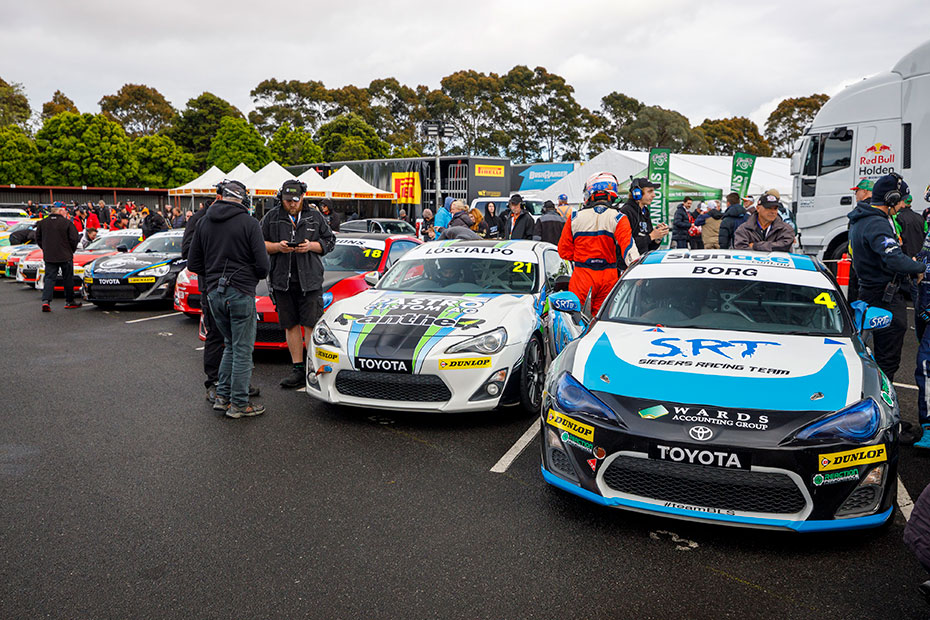 A parking lot full of Toyota G R 86s in racing livery on an overcast day, with racing fans interacting with professional racing drivers.