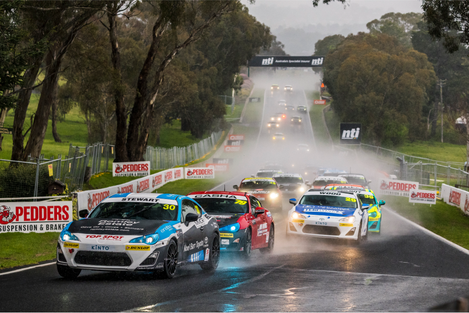 Toyota G R 86s in racing livery compete on a wet race track surrounded by grass and native Australian trees. Watery mist fills the air.