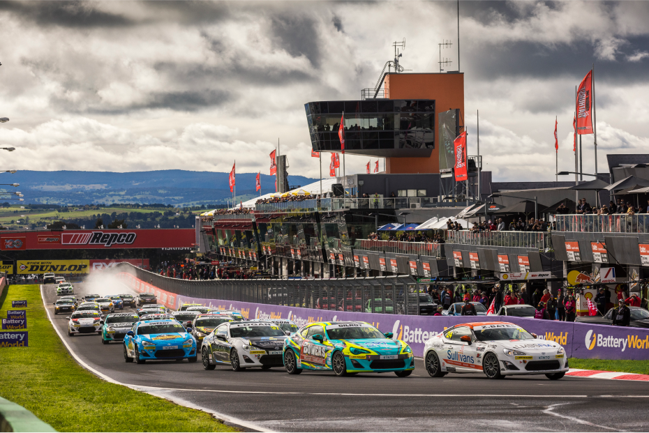 Toyota G R 86s in racing livery compete on a race track lined with grandstands, hundreds of spectators watching. Grey clouds loom above.