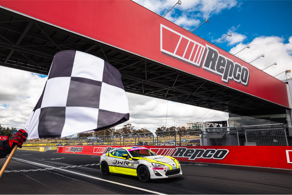A White Liquid Toyota G R 86 in fluorescent yellow racing livery drives across the finish line. A large red sponsor archway hangs overhead.