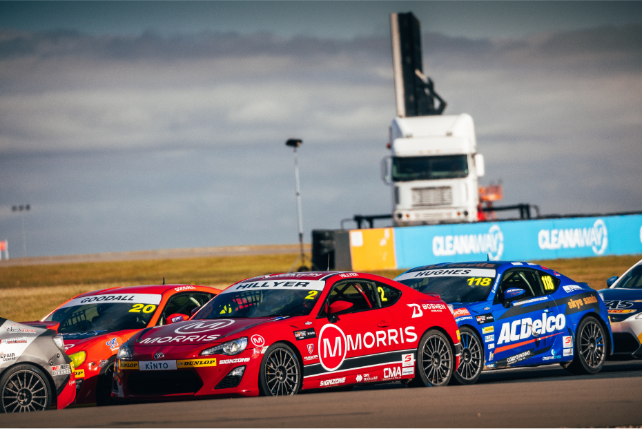 Toyota G R 86s in racing livery competing in a race on an overcast day. A large white truck can be seen in the background.