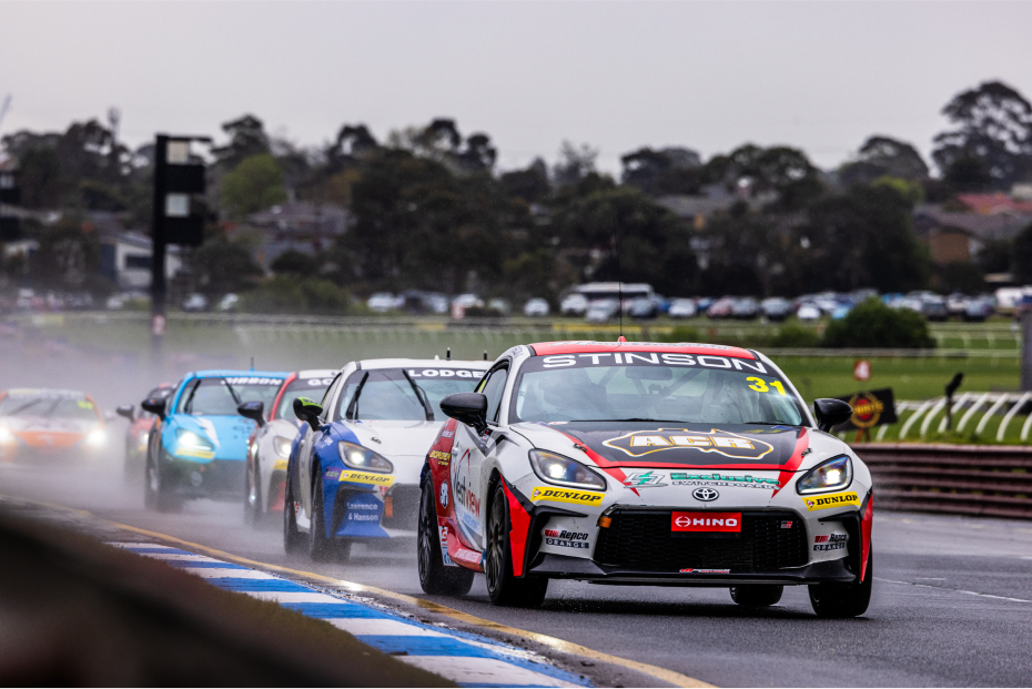 Six Toyota G R 86s competing on a rainy track, some with their headlights on. Trees and housing dot the background.