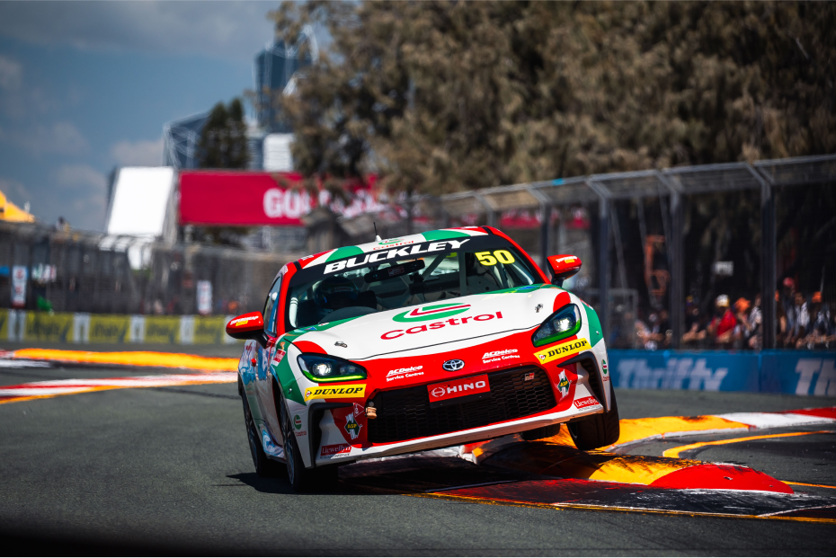 A Toyota G R 86 in red and green racing livery tilted to its right side as it mounts a kerb on a winding race track.