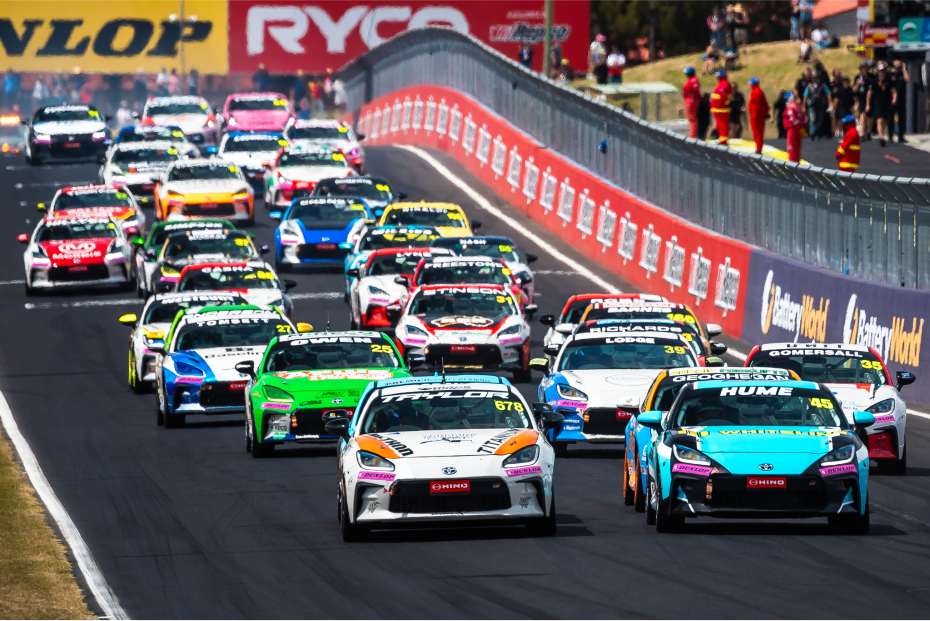A large group of Toyota G R 86s in racing livery competing on a race track. The heat given off by the race is creating a mirage, distorting the vehicles in the distance.