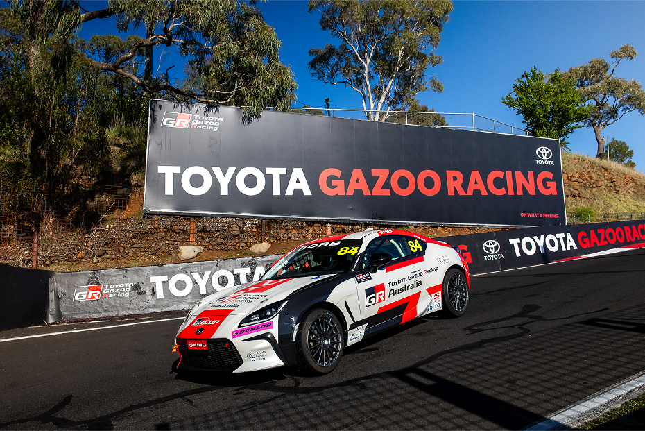 A Toyota GR 86 in red and white racing livery parked on a racetrack incline. A black billboard in the background says 'Toyota Gazoo Racing' in white and red.