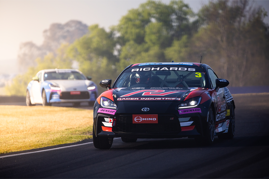 A Toyota G R 86 in Storm Black with red and pink racing livery competing on a race track, a white G R 86 following with headlights on.