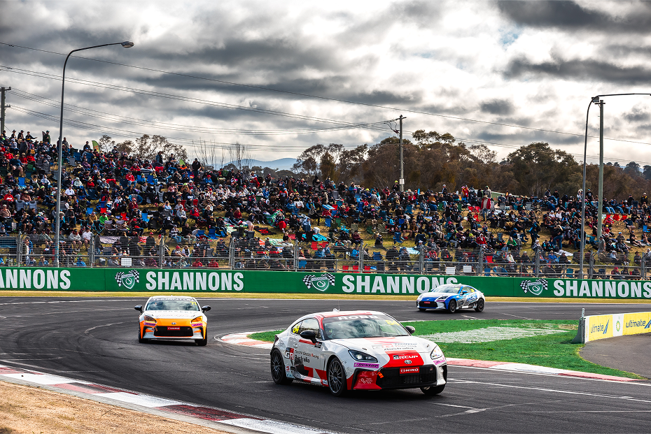 Crowds on an overcast day in the grandstands watch as three Toyota G R 86s in racing livery make the bend on a race track .