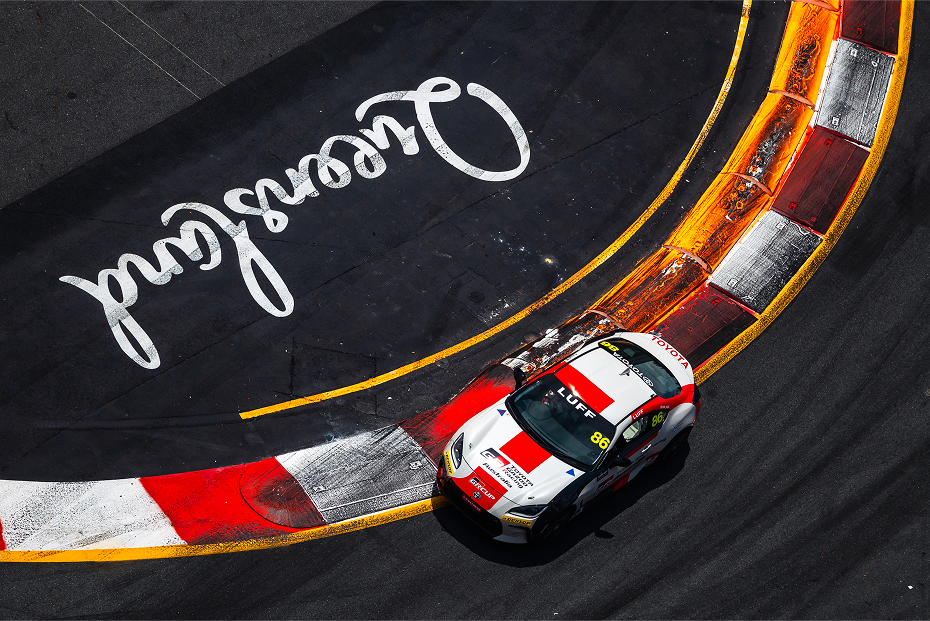 An aerial view of a Toyota White Liquid G R 86 in red racing livery, driving over the inner racetrack kerb as it takes a sharp corner. The word 'Queensland' is painted onto the black inner track in white. 