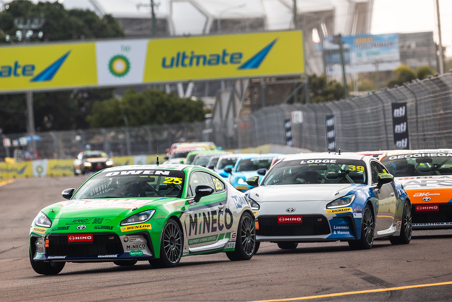 A group of Toyota G R 86s competing on a race track lined with tall wire barriers. A vehicle in green and white racing livery is in the lead.