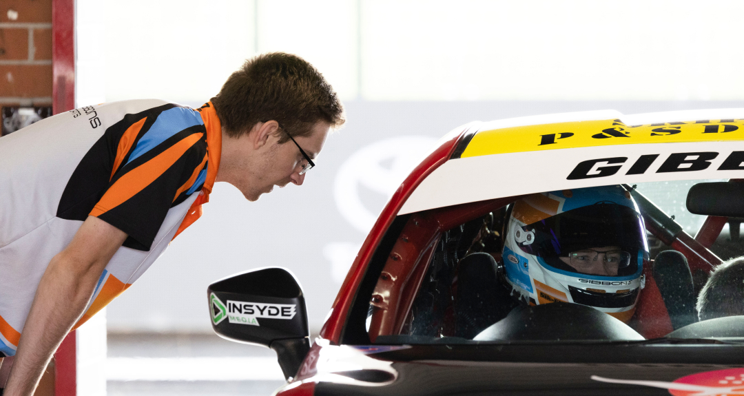 A pit stop mechanic in a blue, black, white and orange polo leans toward the driver's window of a race car to speak to the driver.