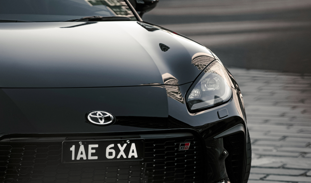 A Toyota G R 86 in Storm Black, the dark grey brick beneath it contrasting with its polished surface. Its black mesh grille features a G R badge.