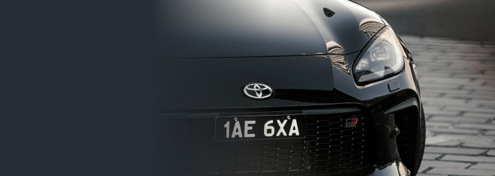 A Toyota G R 86 in Storm Black, the dark grey brick beneath it contrasting with its polished surface. Its black mesh grille features a G R badge.