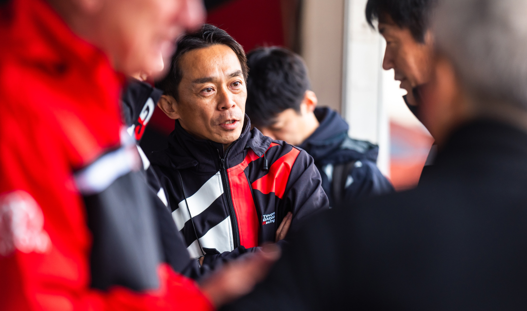 Yuichiro Haruna, the Project Director for the Toyota Gazoo Racing World Rally Team, wearing a black and red T G R jacket, talking to a colleague.
