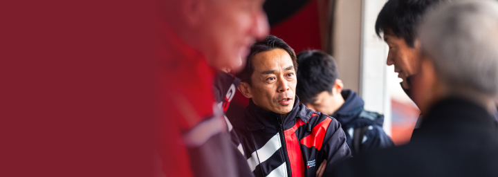 Yuichiro Haruna, the Project Director for the Toyota Gazoo Racing World Rally Team, wearing a black and red T G R jacket, talking to a colleague.
