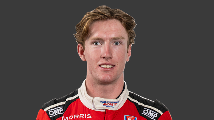 A headshot of professional T G R A driver Matthew Hillyer in a red, navy and white racing suit on a grey background.