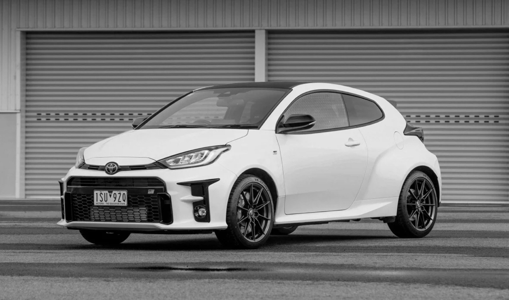 A G R Yaris in Glacier White parked on a wet test track in front of a pair of grey corrugated warehouse doors.