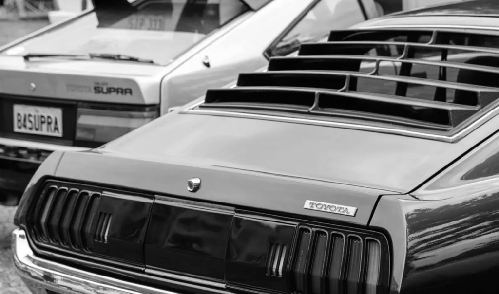 The rear of a black vintage Toyota, its rear windscreen featuring distinct black horizontal louvres. In the background is a silver 1984 Supra slightly out of focus.