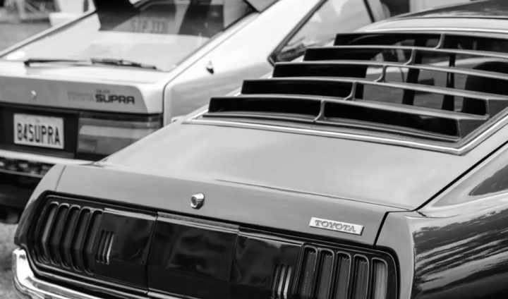 The rear of a black vintage Toyota, its rear windscreen featuring distinct black horizontal louvres. In the background is a silver 1984 Supra slightly out of focus.