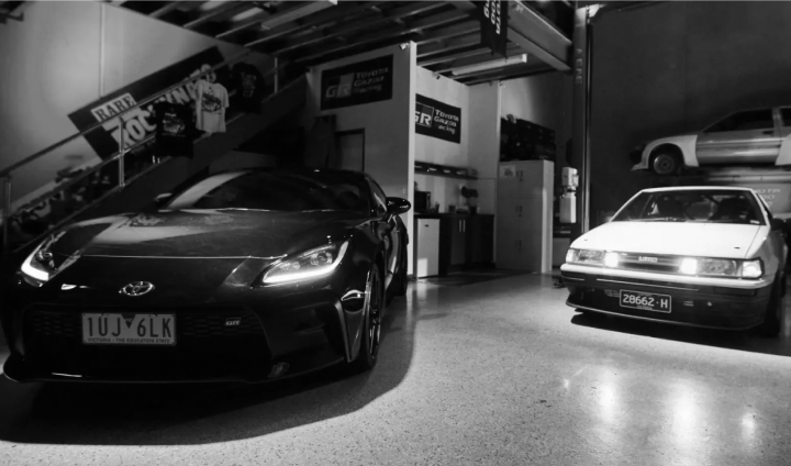 Two Toyota vehicles on display in dramatic red and blue lighting in a warehouse in Melbourne on 86 Day. On the left is a Storm Black G R 86, and on the right is a white Toyota Corolla Levin A E 86.