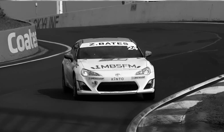 A White Liquid Toyota G R 86 in racing livery on a winding race track. The name 'N.BATES' is above the windshield.