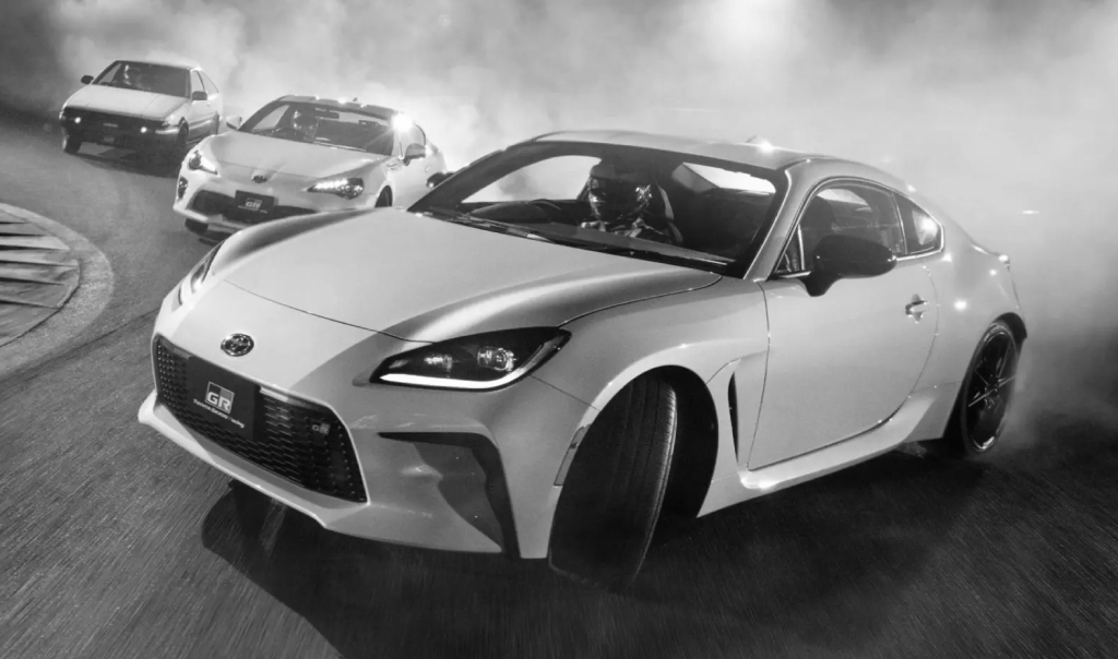 Three generations of the Toyota 86 series in dramatic lighting, with smoke coming from their rear tyres. Closest to furthest: Toyota G R 86, Toyota 86, Toyota A E 86.