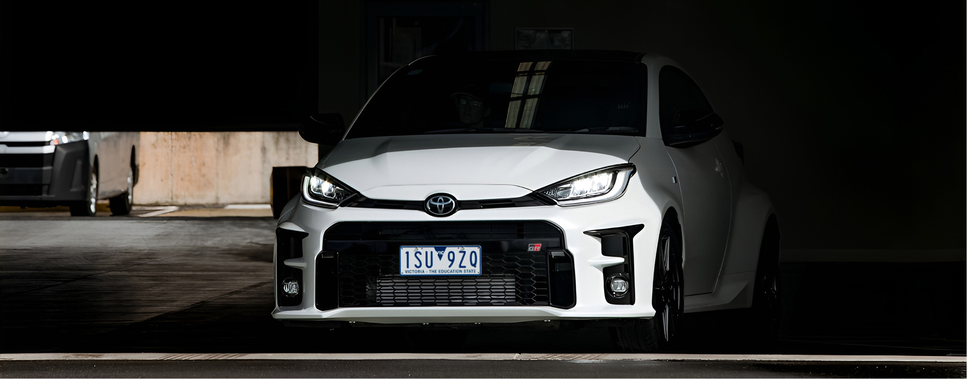 A Glacier White Toyota G R Yaris parked in a pitch-black garage, its nose peeking out into the daylight.