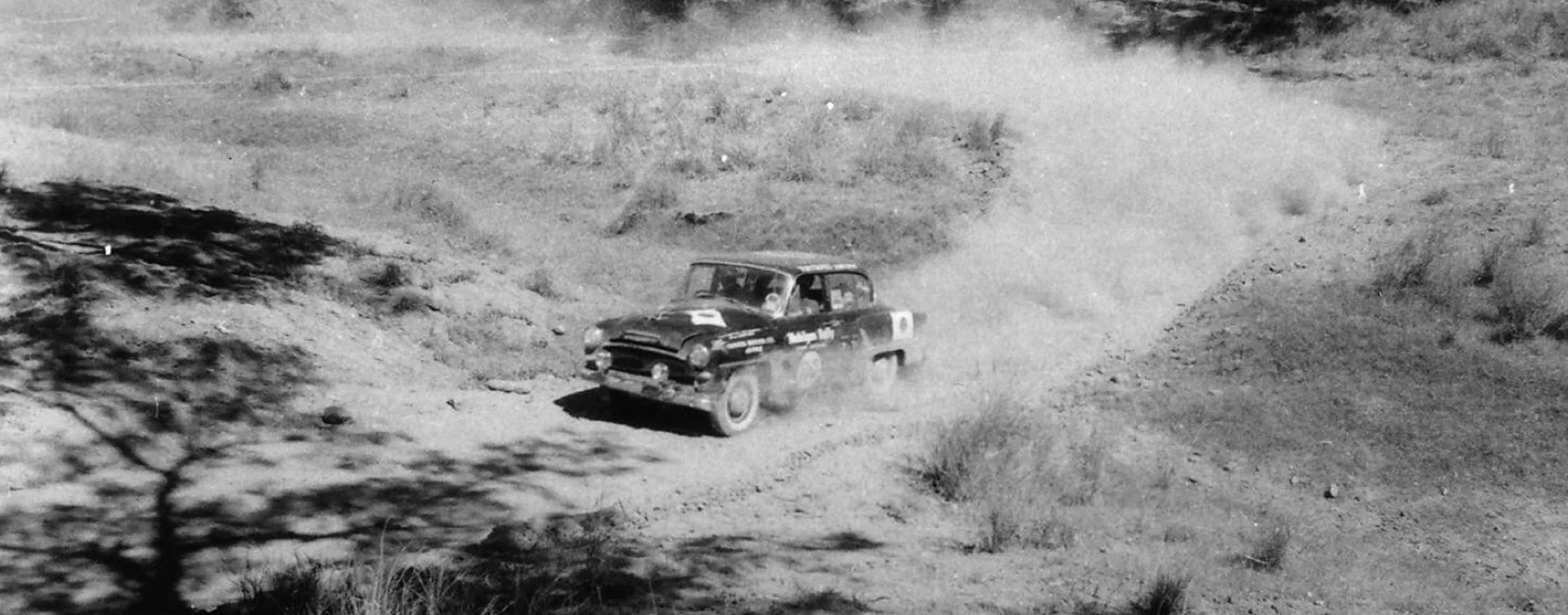 A black and white photograph of a Toyopet Crown R S D taking part in the 1957 Mobilgas Round Australia Rally. Plumes of dust are left in the vehicle's wake as it drives through the Australian bush.