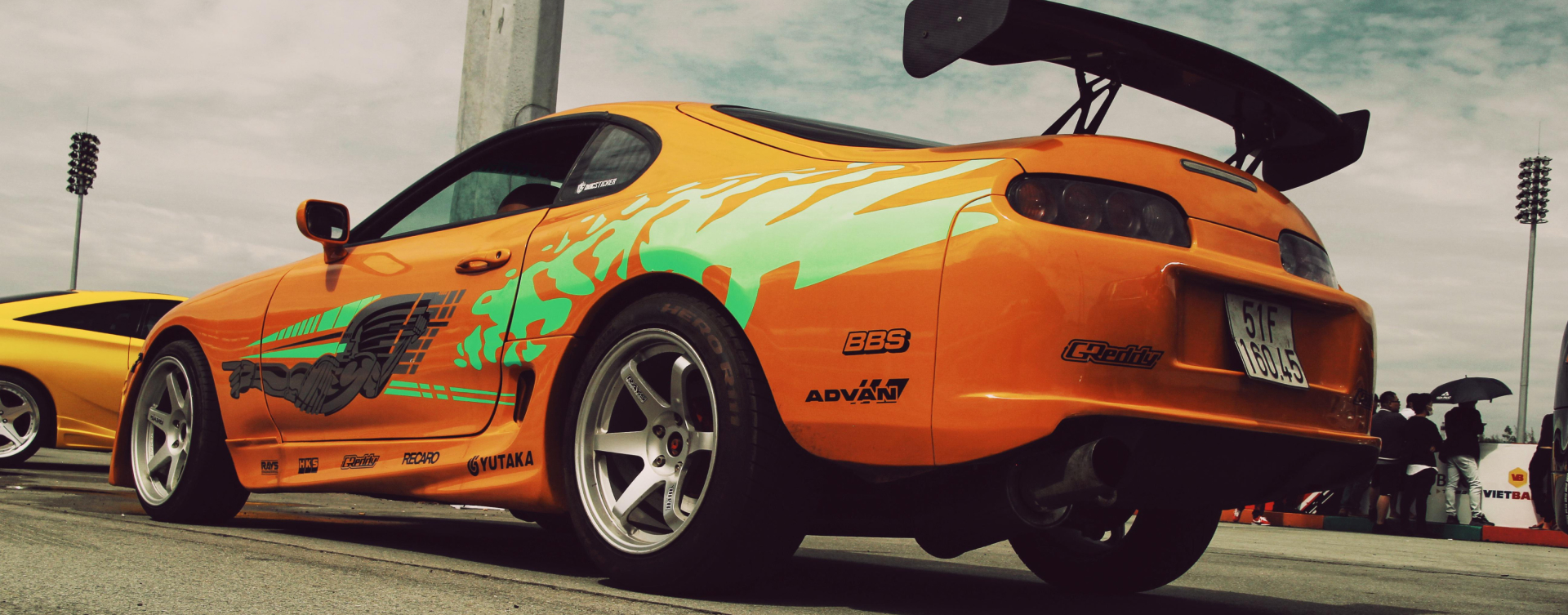 A rear side-view of an orange Toyota Supra Mark 4, with grey and neon green graphic decal. Smaller brand decals run along the bottom.