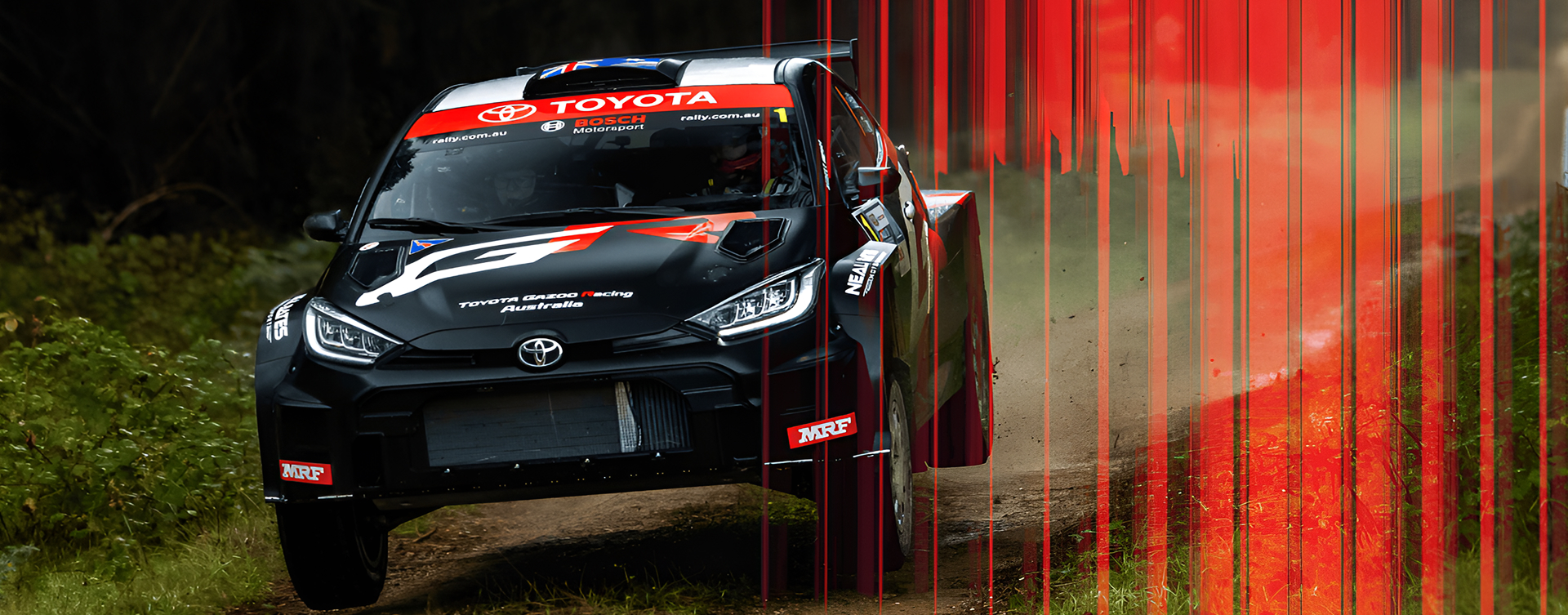 A Tarmac Black Toyota G R Yaris Rally2, midair as it drives over an uneven dirt rally track in a forested area. It’s covered in rally sponsor livery.