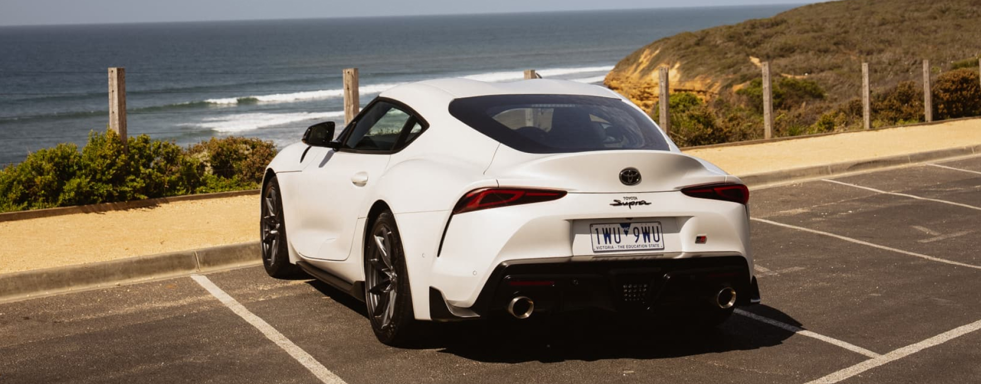 A rear three-quarter view of a white Toyota Supra parked in a coastal lot, with ocean waves and cliffs in the background.