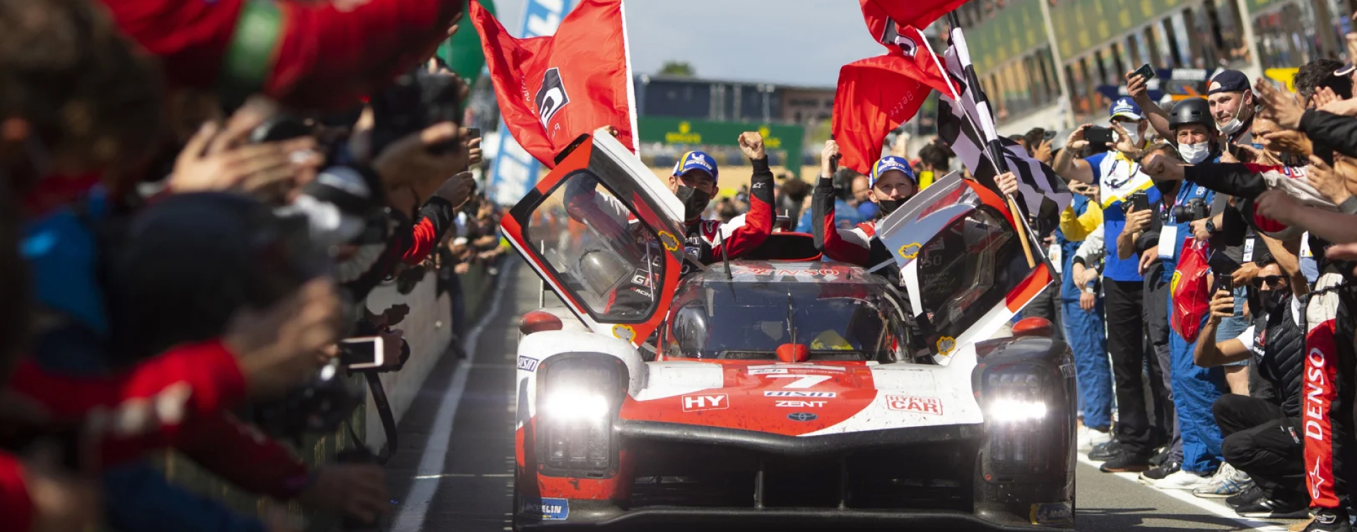 TOYOTA GAZOO Racing Drivers in the number 7 G R 010 HYBRID, celebrating their victory at Le Mans 24 Hours at the Circuit de la Sarthe. The doors of the hypercar are open as it rolls by trackside motorsports fans, with each driver waving a red TOYOTA GAZOO Racing flag.