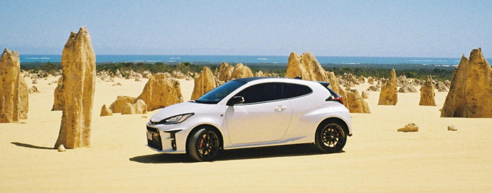 A Toyota G R Yaris Rallye in Glacier White among sharp limestone formations at the Pinnacles Desert. The sleek white vehicle contrasts with its rugged surroundings.