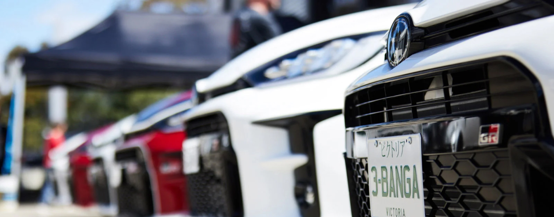 A low-angle view of G R Yaris vehicles lined up into the distance. The nearest G R Yaris in Glacier White is in focus, with its honeycomb grille, 'G R' badge and Japanese-style number plate.