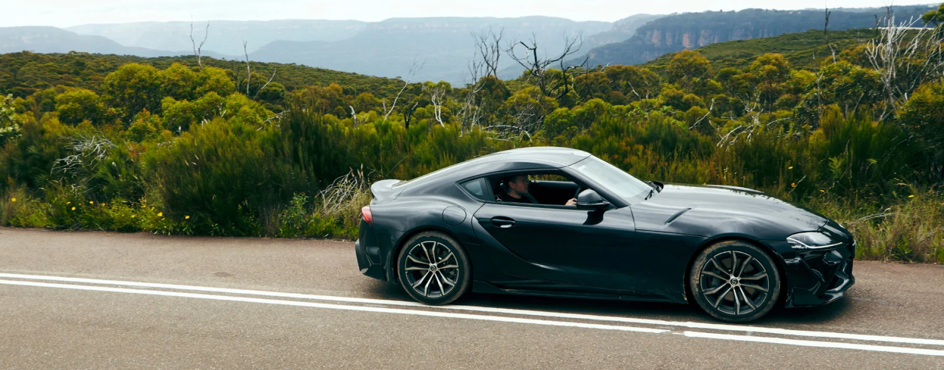 A G R Supra in Bathurst Black on a highway near the Blue Mountains. Native Australian flora stretches into the distance.