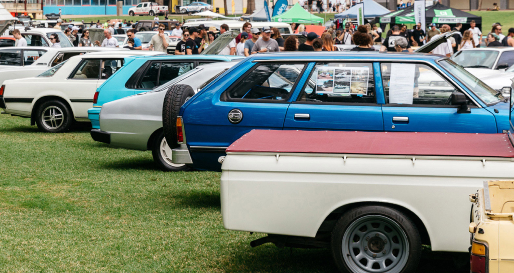 Crowds of people attending Toyota's 'All Japan Day' 2022 in Adelaide. A range of Japanese car makers from across the decades sit on display, parked in rows on a green lawn. Stall tents are pitched in the distance.