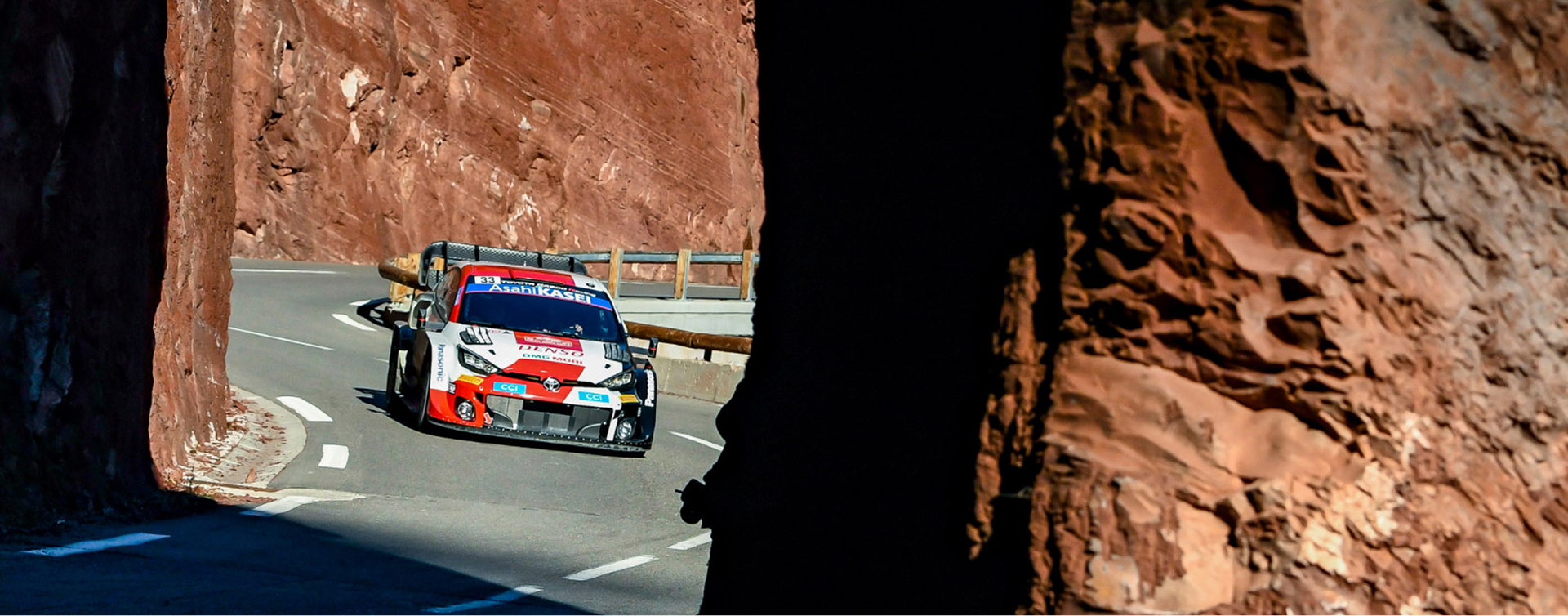 A Glacier White Toyota G R Yaris Rally 1, driving along a road that winds around a rocky red cliff. The vehicle is covered in livery featuring various racing sponsors.