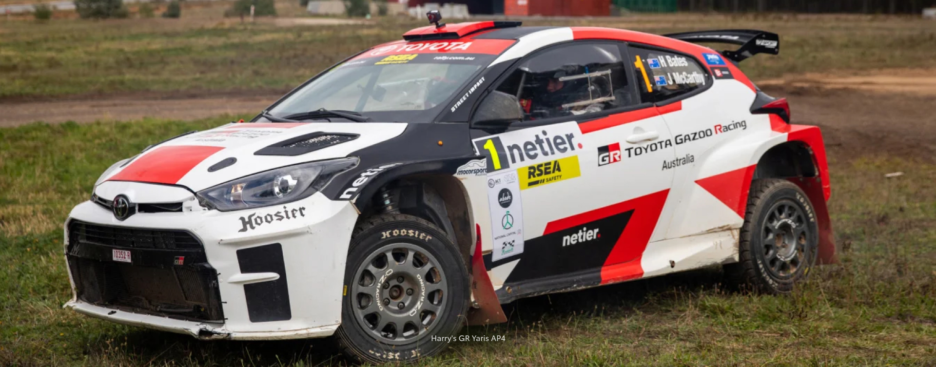 Professional rally driver Harry Bates in the driver's seat of a Glacier White Toyota G R Yaris A P 4. The vehicle is covered in red and black racing livery, featuring TOYOTA GAZOO Racing Australia, as well as various rally sponsors.