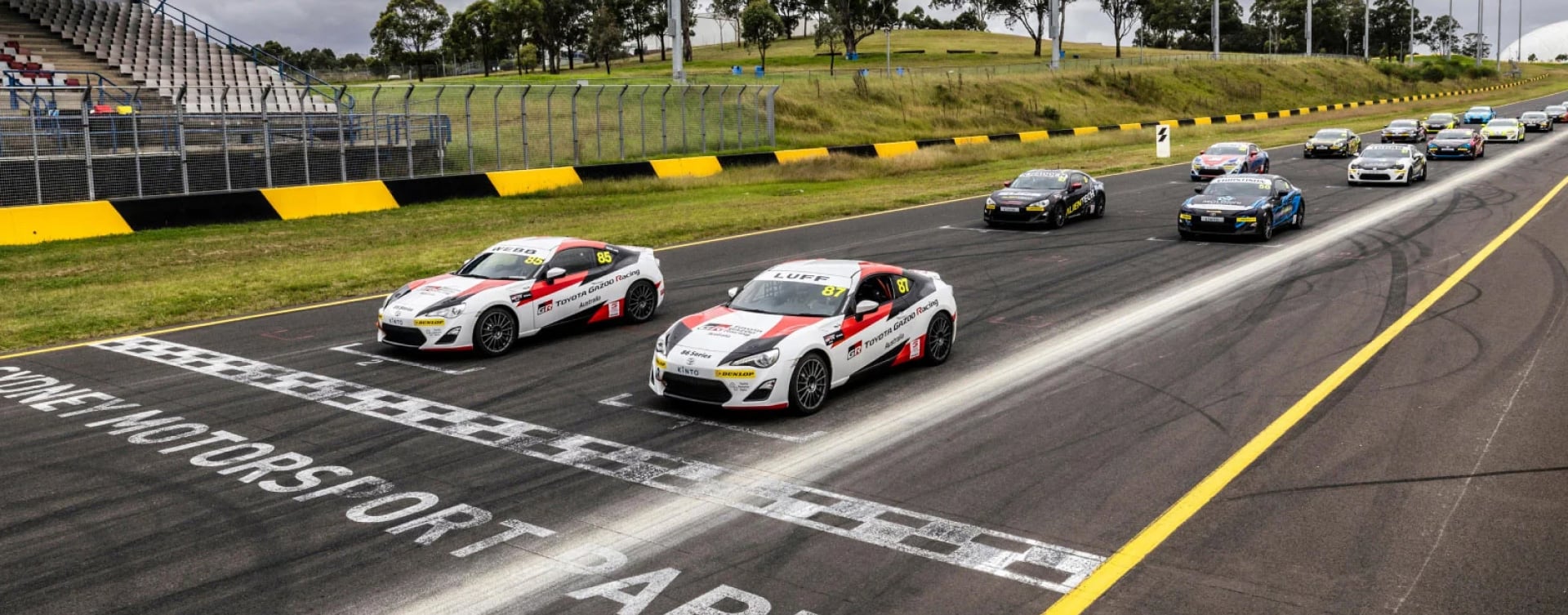 A fleet of 14 Toyota G R 86s in racing livery, at pole position at Eastern Creek raceway, ready to burn some rubber on the track.