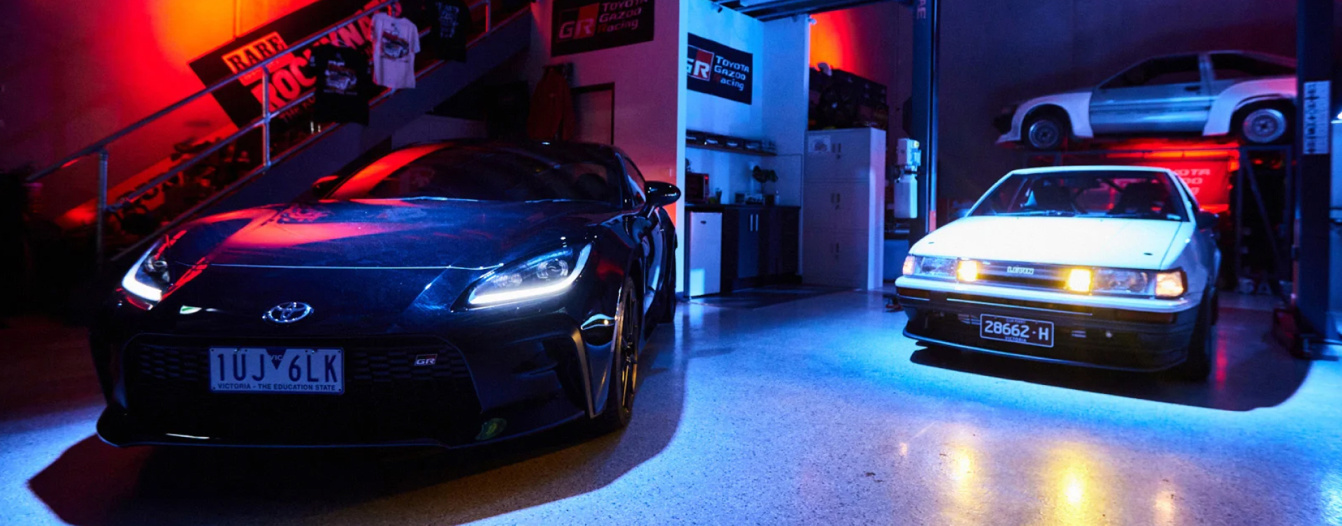 Two Toyota vehicles on display in dramatic red and blue lighting in a warehouse in Melbourne on 86 Day. On the left is a Storm Black G R 86, and on the right is a white Toyota Corolla Levin A E 86.