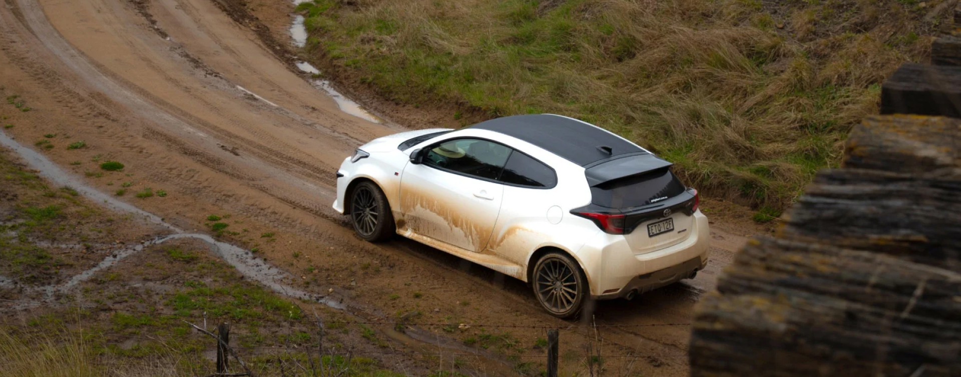 Aerial view of a muddy Toyota G R Yaris in Glacier White driving up a winding, wet road in rural Australia.