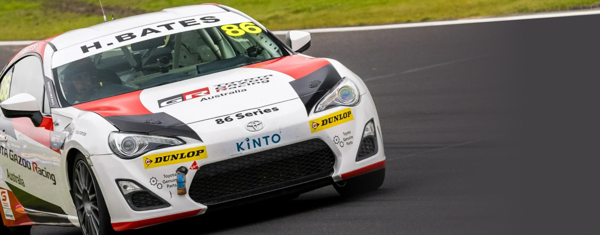 A white Toyota G R 86 in livery on a race track. The name 'N.BATES' is above the windshield. Dramatic angle.
