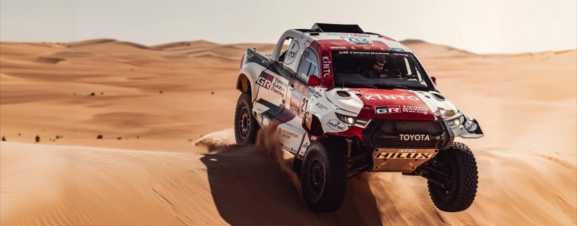 A Toyota Dakar HiLux in red and white T G R livery, front wheels mid-air as it drives over a large orange dune. Desert stretches into the distance.