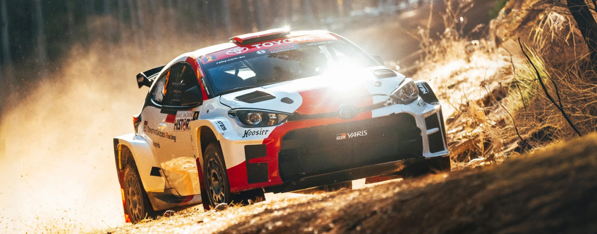 A Glacier White G R Yaris A P 4 in sponsor livery on a dirt rally tack, glistening in the sun. Angular shot.